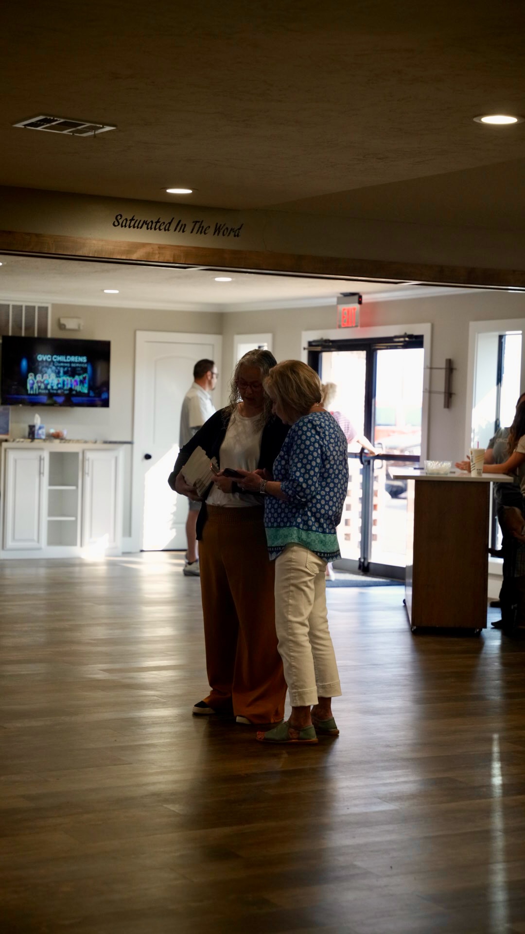 Two women talking under the 'Saturated In The Word' sign